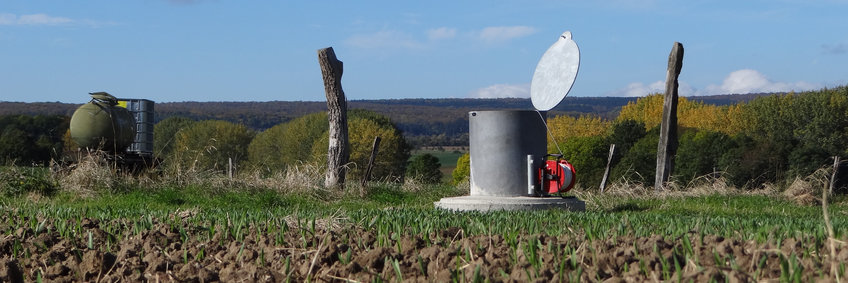 © R. Lehmann, Universität Jena/Hydrogeologie At the edge of a field, a gray cylinder with an open lid stands out of the ground.