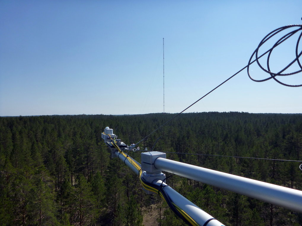 Im Vordergrund ein Strahlungsausleger aus Aluminium mit verschiedenen Straghlungssensoren am Eddyturm in Zotino. Der  Blick zeigt über die dunkelgrüne Taiga zum 300 m hohen ZOTTO-Turm in in wenige Kilometer Entfernung.
© M. Hertel/BGC