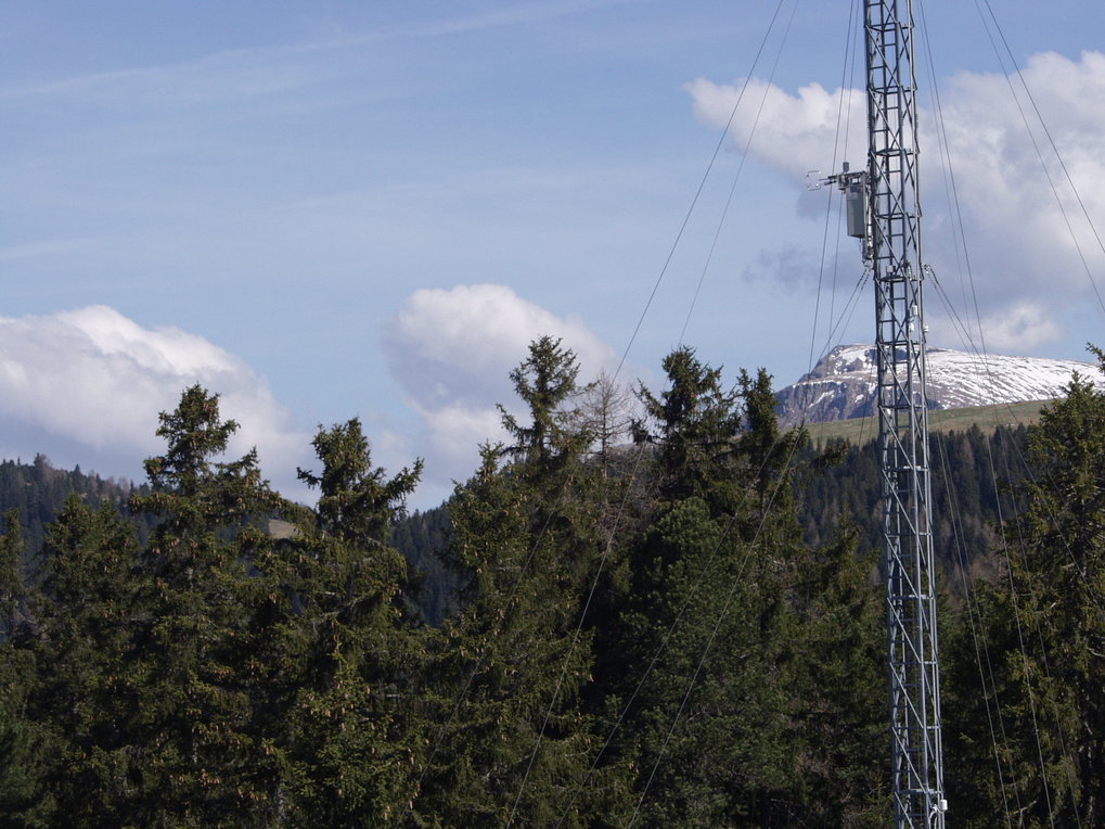 Eddy-Corarianz-Turm in Renon - Italien, während des Advex-Experimentes. Teil eines Gittermast am rechten Bildrand mit etlichen ABspannungen. vor einem Fichtenwald im Gebirge. Ein Schneebedeckter Berg im Hintergrund. Gro0e weisse Wolken am hellblaverschleiertem Himmel.