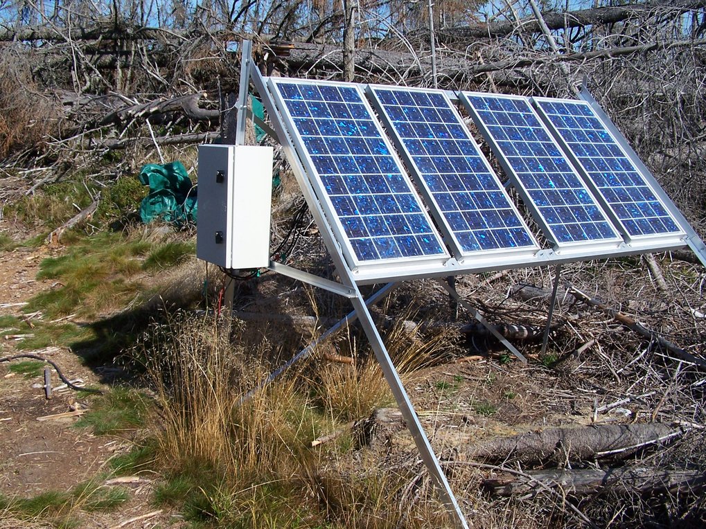 Sonnebeschienene Solaranlage mit Schaltschrank, hier 4 blaue Solarpanel auf einem Alugerüst im Windbruch. Überall totes graues Holz der umgeworfenenen  Nadelbäume, dazwischen ein paar Gräser.
© MHertel/BGC