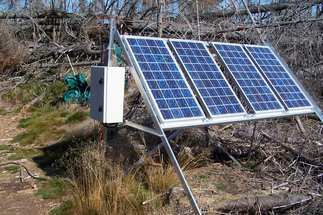 Sonnebeschienene Solaranlage mit Schaltschrank, hier 4 blaue Solarpanel auf einem Alugerüst im Windbruch. Überall totes graues Holz der umgeworfenenen  Nadelbäume, dazwischen ein paar Gräser.
© MHertel/BGC