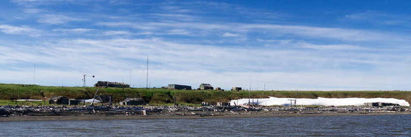 View of the Ambarchik station from the sea (© M. Hertel/BGC) View of the Ambarchik station from the sea, several buildings and masts in the distance, green meadows, blue sky with white cirrus clouds, a larger amount of snow is still on the beach, there is also a lot of gray driftwood with a few wooden huts between them