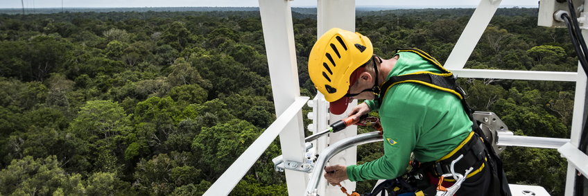 In the foreground, Max Planck employee Kerstin Hippler is seen attaching a Söll fall protection rail; she is wearing a yellow climbing helmet over a red baseball cap and a fall protection harness over an LA shirt in Brazilian green. She works secured at a height of about 100m on the tower, which is 300m high in total. Around her is the white painted scaffolding structure and in the background you can see the tropical rainforest to the horizon.