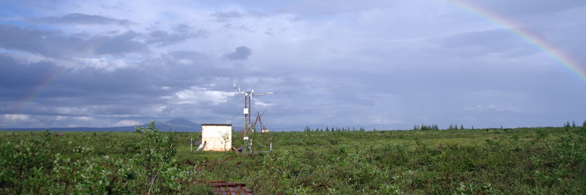 You can see a small hut with a measuring tower, which is equipped with a variety of sensors. One looks over the drained area in the flood plain in the Kolyma lowlands. 
It is summer. In the foreground a few small trees, otherwise the landscape is green and flat until the horizon, where you can see a mountain cone. The sky is full of clouds and a rainbow stretches from left to right across the entire sky.