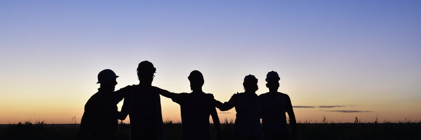 Group picture of Brazilian, American and German scientists in front of setting sun. You can see only the silhouette of 5 people who put their arms on and around each other's shoulders, in front of a magical sunset. The sky shows a color gradient from purple-blue to golden-orange.