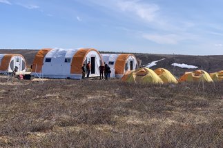Camp in der Tundra Kanadas, die nächste größere Stadt ist Inuvik am Yukon.
Auch hier ist es im Sommer 22 viel zu warm. Man sieht eine braune Graslandschaft vor strahlend blauem Himmel. Links im Bild stehen 3 große Tonnenzelte in orange und weiß und rechts davon bzw. davor stehen 5 kleinere geodätische Zelte in gelb und orange. Das Camp ist von einem  Weidezaun umgeben und in der Mitte ragt eine kleine Wettterstation auf. Im Hintergrund sieht man noch ein paar kleine Ecken mit Schneeresten.