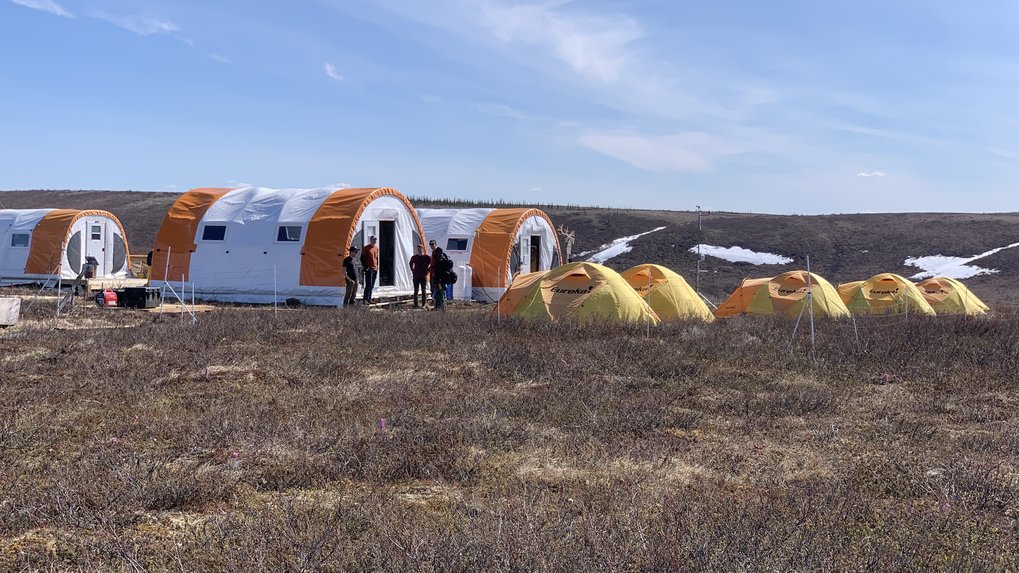 Camp in the tundra of Canada, the nearest major city is Inuvik on the Yukon.
Here, too, it is much too warm in summer 22. You see a brown grassy landscape in front of a bright blue sky. To the left of the picture are 3 large barrel tents in orange and white and to the right of them or in front of them are 5 smaller geodesic tents in yellow and orange. The camp is surrounded by a pasture fence and in the middle there is a small betting station. In the background you can see a few small corners with snow remains.