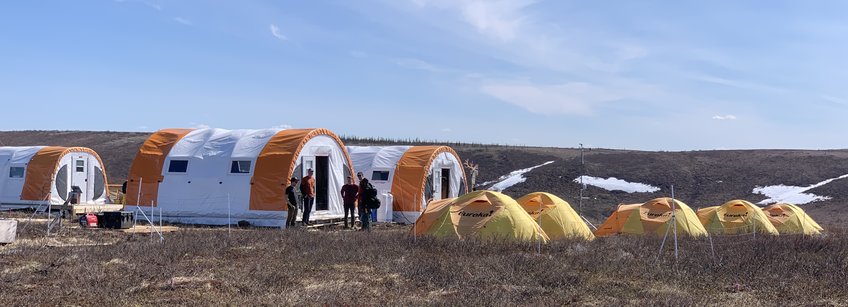 Camp in the tundra of Canada, the nearest major city is Inuvik on the Yukon.
Here, too, it is much too warm in summer 22. You see a brown grassy landscape in front of a bright blue sky. To the left of the picture are 3 large barrel tents in orange and white and to the right of them or in front of them are 5 smaller geodesic tents in yellow and orange. The camp is surrounded by a pasture fence and in the middle there is a small betting station. In the background you can see a few small corners with snow remains.