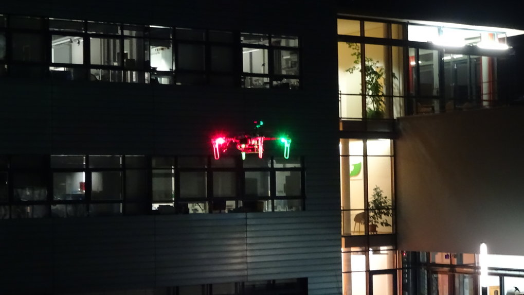 Im Hintergrund dass teilweise beleuchtete Institutsgebäude, im Vordergrund eine farbig leuchtende Forschungsdrohne.