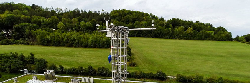 Top of the meterology mast of the MPI-BGC in Jena, with various measuring instruments for radiation, temperature, wind and humidity. In the background, the green meadows of the Beutenberg and leafy trees on the Ammerbacher Platte.