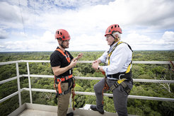 Max Planck researcher Stefan Wolff and German President Steinmeier stand on the railing of Atto's first platform. They are wearing climbing helmets and harnesses. In the background, the rainforest stretches to the horizon.