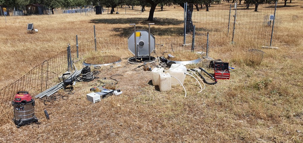 Lysimeterwartung in Majadas (Spanien). Technisches Gerät, Kanister und Staubsauger auf einer verbrannten Wiese.
Vegetation in einem silbernen Ring, daneben eine geöffnete Klappe, die in die unteridische Einrichtung des Lysimeters führt. Darum herum ein kleiner Drahtzaun, der die Weidetiere hindern soll, die Messfläche zu betreten.