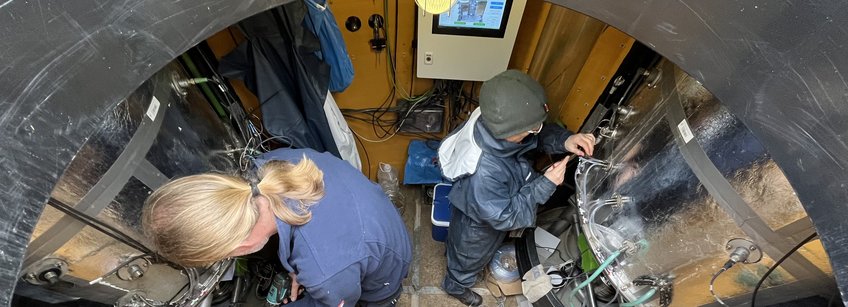 Inside a lysimeter of the company UGT in Majadas Spain. Two people repairing/maintaining the system. Two large insulated columns on the right and on the left in the picture above a formwork cabinet with visual button and at the bottom in the picture the ladder stairs. In between the orange plastic walls of the inner lining of the lysimeter cellar.