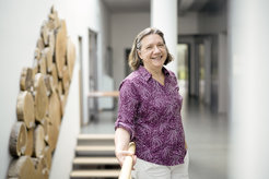 Woman in a purple shirt stands leaning diagonally against stair railings, in the background a multitude of tree slices as decoration on the wall.