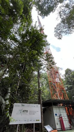 View from the ground up to the 325 metre high Atto Tower. In the foreground on the left, a light-coloured board with all the cooperation partners involved printed on it. To the right is a covered container. And from the bottom right to the top centre of the portrait format image is the scaffolding tower, which is painted alternately orange and white every 12 metres. There is a stairwell in the tower with an accompanying safety rail (INNOTECH) and the tower is hollow in the centre with a rail for a one-person lift. There is also a lift rail on the corner spar facing the tower on which a robot lift regularly measures profiles on a scientific mission. The tower is surrounded by trees up to 40 metres high. The sky is mostly white in the photo, interspersed with a few patches of blue.