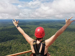Eine Person mit rotem Kletterhelm steht auf einer Plattform und blickt über einen ausgedehnten, grünen Regenwald, mit erhobenen Armen in einer Geste des Jubels.