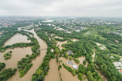 Luftaufnahme einer überschwemmten Stadtlandschaft mit Bäumen, Brücken und Gebäuden im Hintergrund.