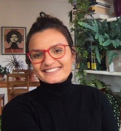 Portrait einer lächelnden Frau mit dunklen Haaren, schwarzem Rollkragenpullover und roter Brille, im Hintergrund ein Bücherregal mit vielen Pflanzen.