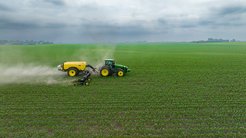 A green tractor with yellow wheels moves across a large agricultural field, spraying crops and leaving a visible dust trail behind under a cloudy sky.