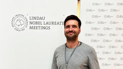 Eine Person in einem grauen Oberteil steht vor einer Wand, die mit dem Logo der Lindau Nobel Laureate Meetings dekoriert ist, das die Worte "Educate. Inspire. Connect." enthält.