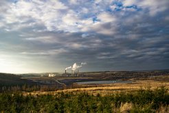A power plant with multiple smokestacks emits steam in a vast rural setting, surrounded by fields and forests under a cloudy sky.