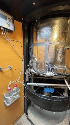 Underground measuring device in an orange roundel manufactured by the company UGT. You can see a large silver-clad barrel with a diameter of approx. 1 meter and a height of 1.5 meters standing on a weighing frame. A number of hoses and cables come out of the barrel, which are connected to tensiometers and suction cups. Inside the weighing frame are pumps and a stainless steel water tank.