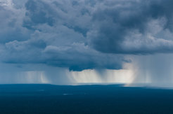 Dramatische, dunkle Regenwolken erstrecken sich über einen bewaldeten Horizont, während im Hintergrund starker Regen fällt