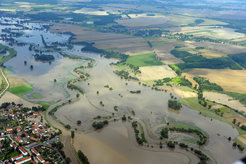 Luftaufnahme eines Flusses, der über die Ufer getreten ist und eine Auenlandschaft überschwemmt, aber auch eine Siedlung in der linken unteren Bildecke erreicht hat. Der Fluss nimmt den Teil des Bildes von linken oberen bis zu rechten unteren Ecke ein. Im Hintergrund ist eine Landschaft mit von kleinen Wäldern, einzelnen Bäumen und Büschen durchbrochenen Feldern zu sehen.